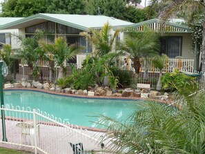 A pool area with a fenced deck, palm trees, and a building with a green roof. - Kangerong Holiday Park (Dromana)