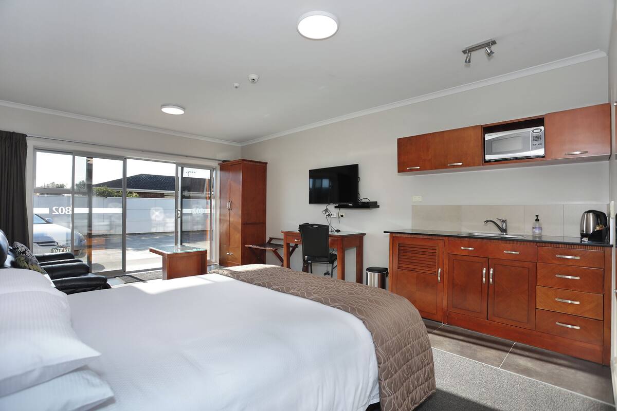 A hotel room with a large bed, a desk, a chair, a television, and a kitchenette.