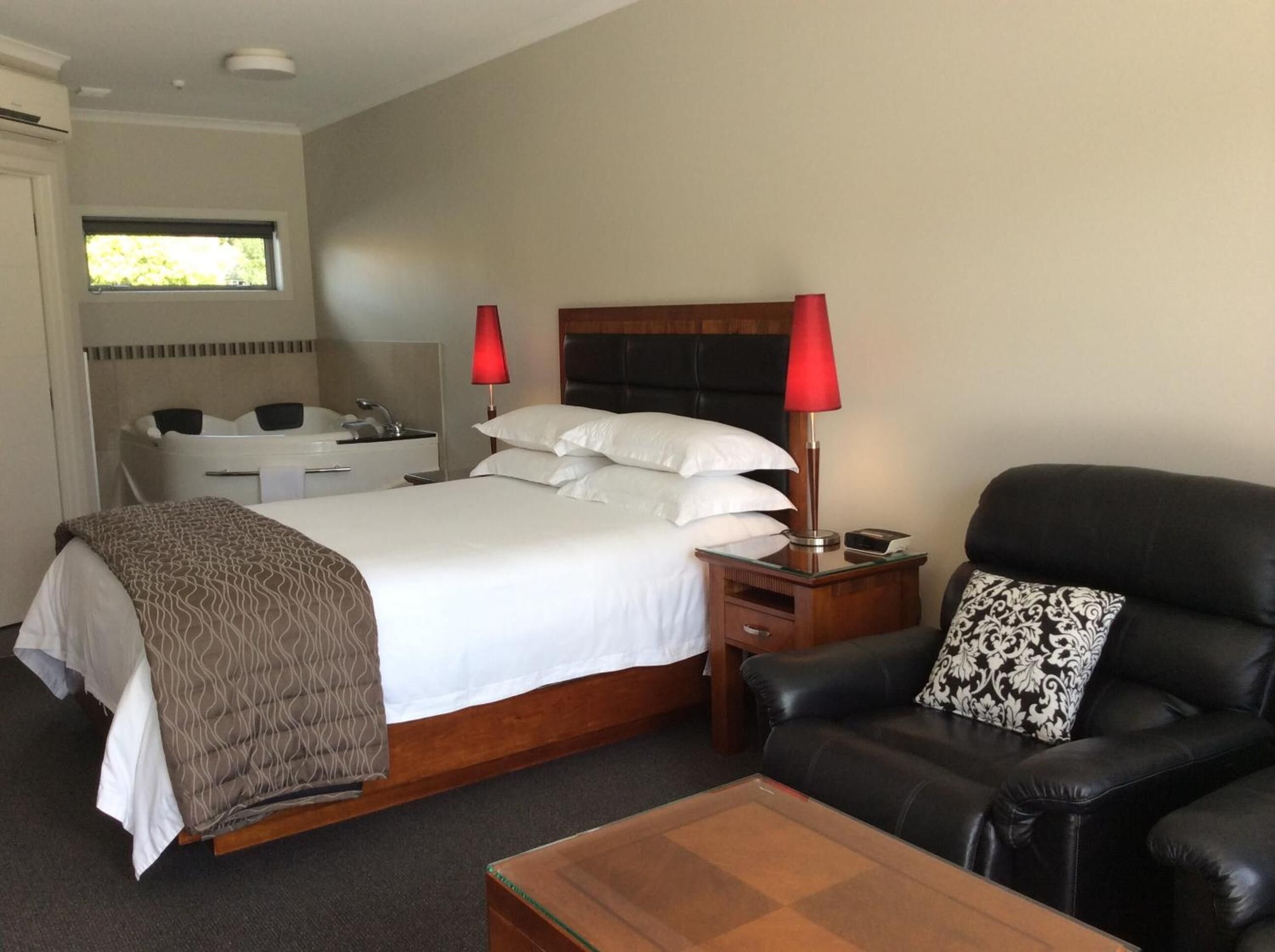 Una habitación de hotel con cama, mesita de noche, sillón de cuero negro, mesa de centro de vidrio y baño con bañera y lavamanos.