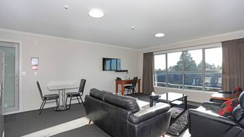 A modern living room with a sofa, chairs, a coffee table, and a TV. There is a dining table with chairs, a window with curtains, and a door.