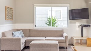 A living room with a beige sectional sofa, a matching ottoman, a flat-screen TV mounted on the wall, and a window with a view of a plant outside. - NRMA Portland Bay Holiday Park (Portland)