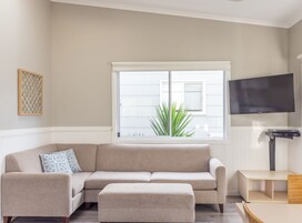 Una sala de estar con un sofá seccional color beige, un reposapiés a juego, un televisor de pantalla plana montado en la pared y un ventanal con vista a un jardín.