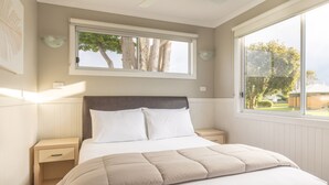 A bedroom with a bed, nightstands, a window, and a view of trees and buildings. - NRMA Portland Bay Holiday Park (Portland)