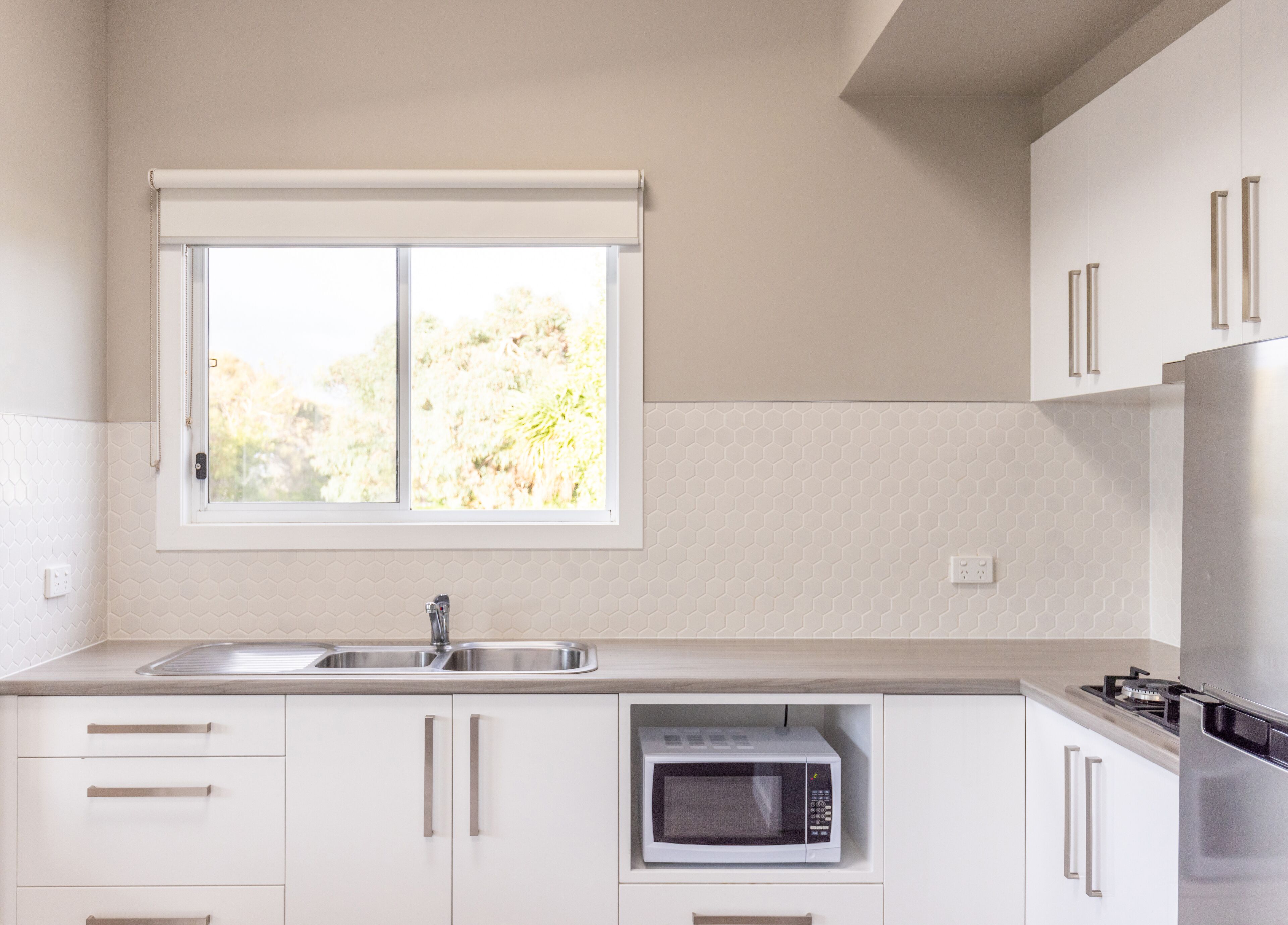 A modern kitchen with white cabinetry, a stainless steel sink, a built-in microwave, and a window with a view of trees.