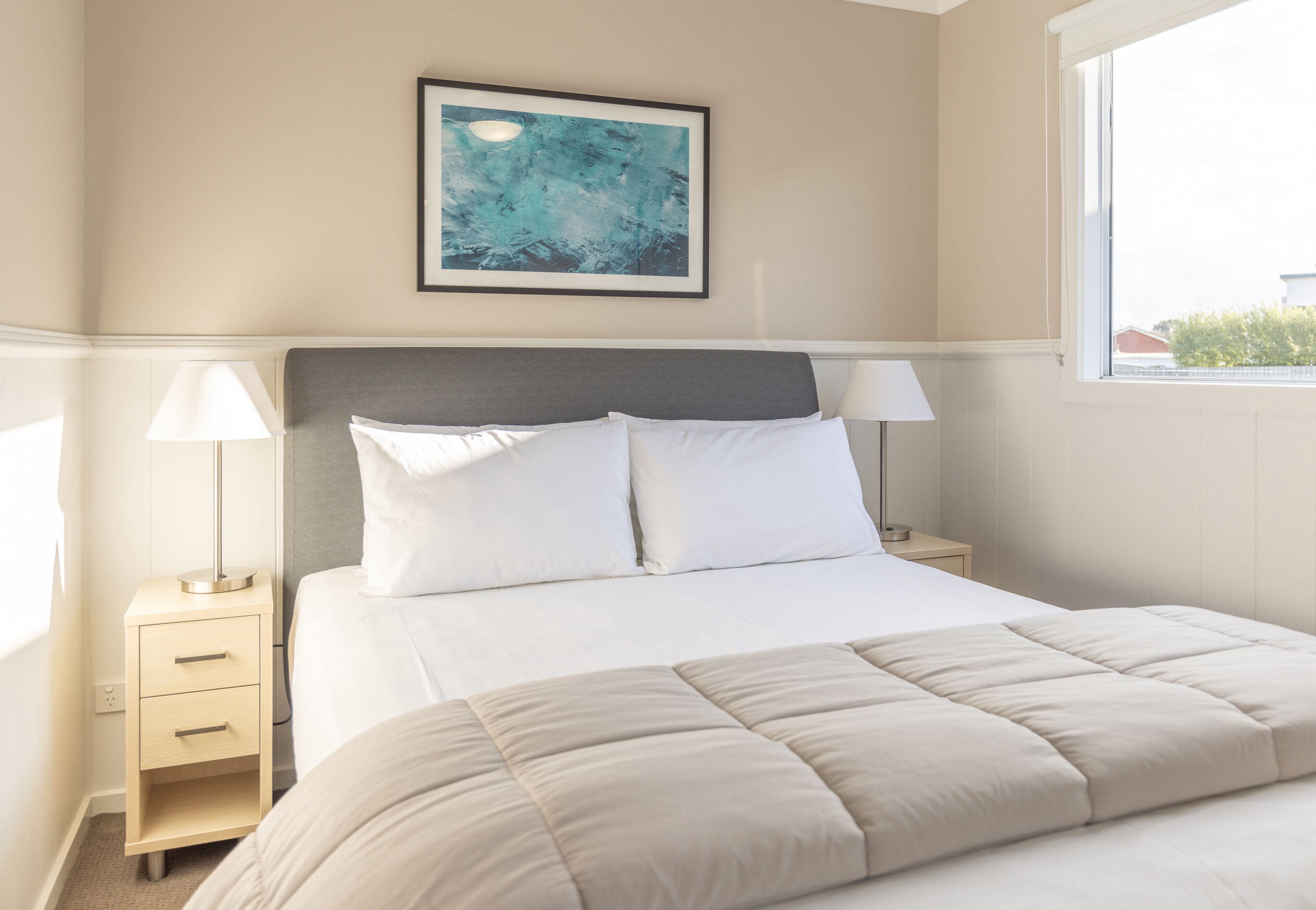Cama bem arrumada com cabeceira cinza, duas mesas de cabeceira com abajures, um quadro abstrato emoldurado na parede e uma janela com vista para prédios.