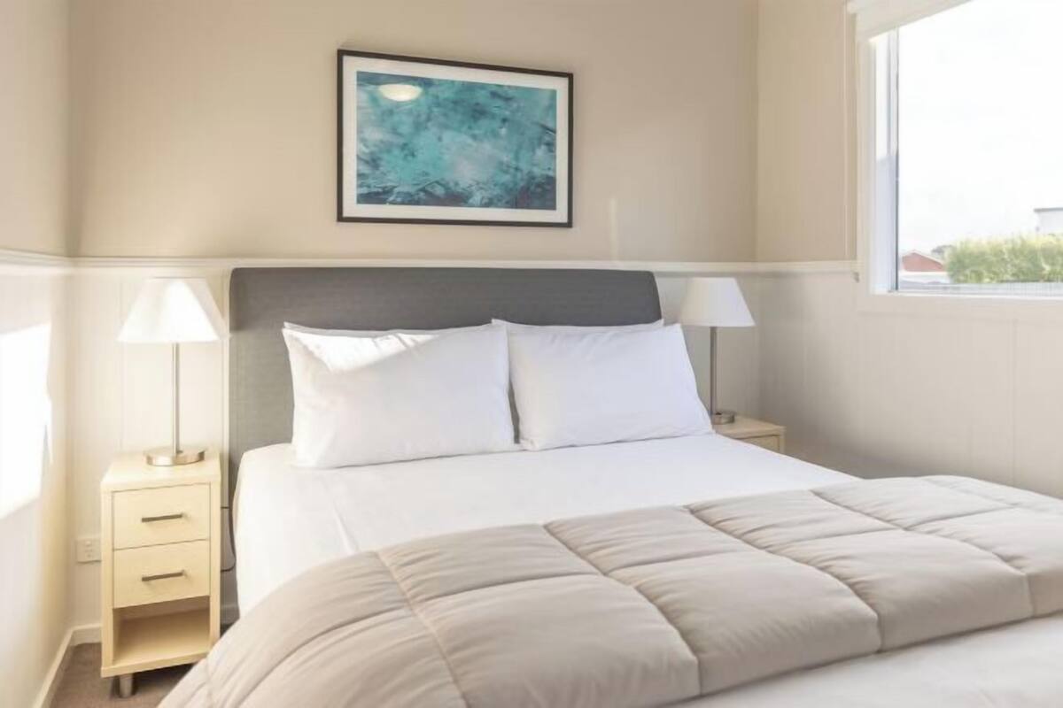 A hotel room with a bed, two bedside tables, a nightstand, a framed picture on the wall, and a window.