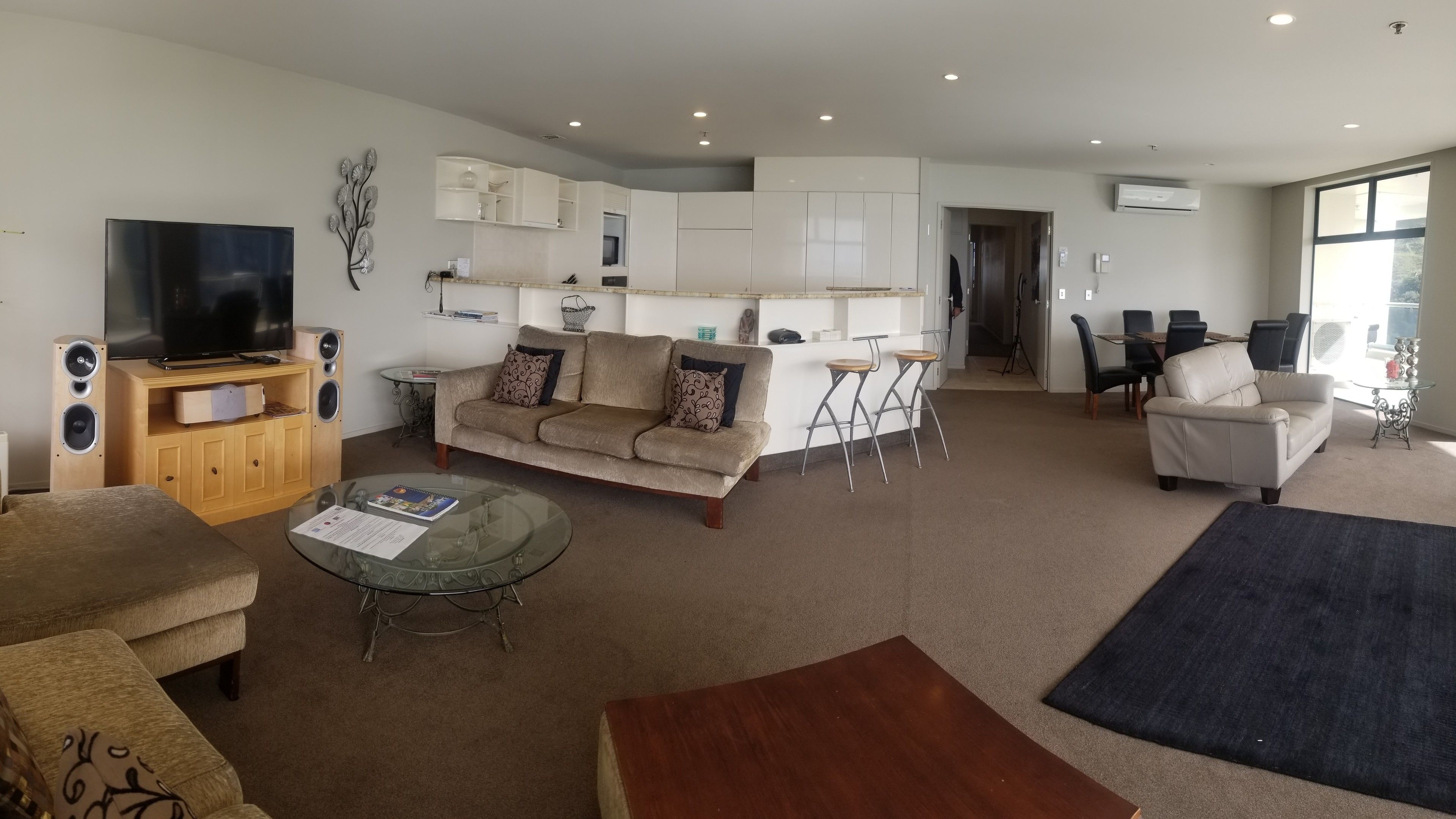 A modern living room with a sofa, coffee table, and TV. There is a kitchen area with cabinets and a dining table.