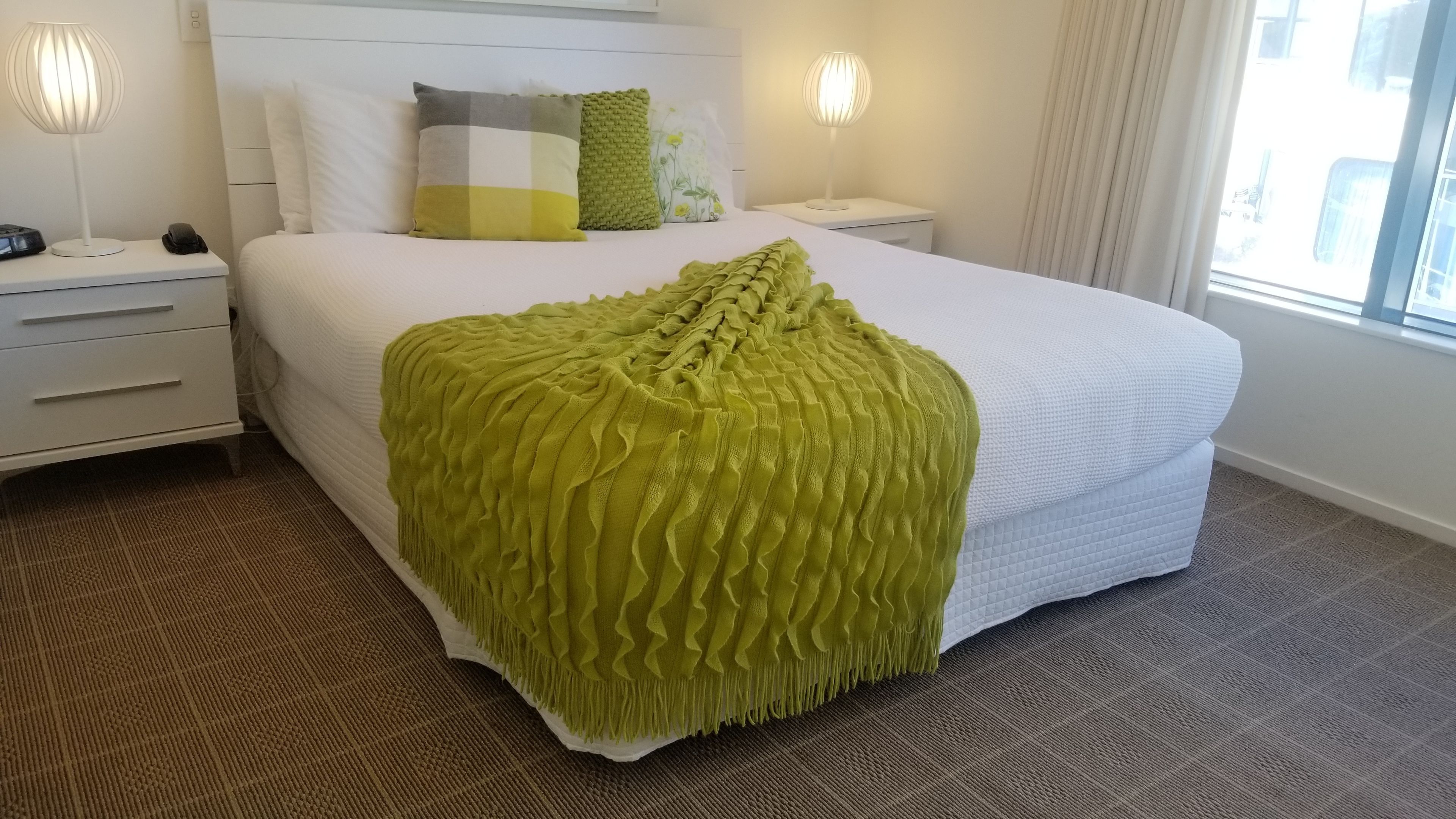 A neatly made bed with a green blanket, a bedside table with a lamp, a phone, and a nightstand with a lamp.