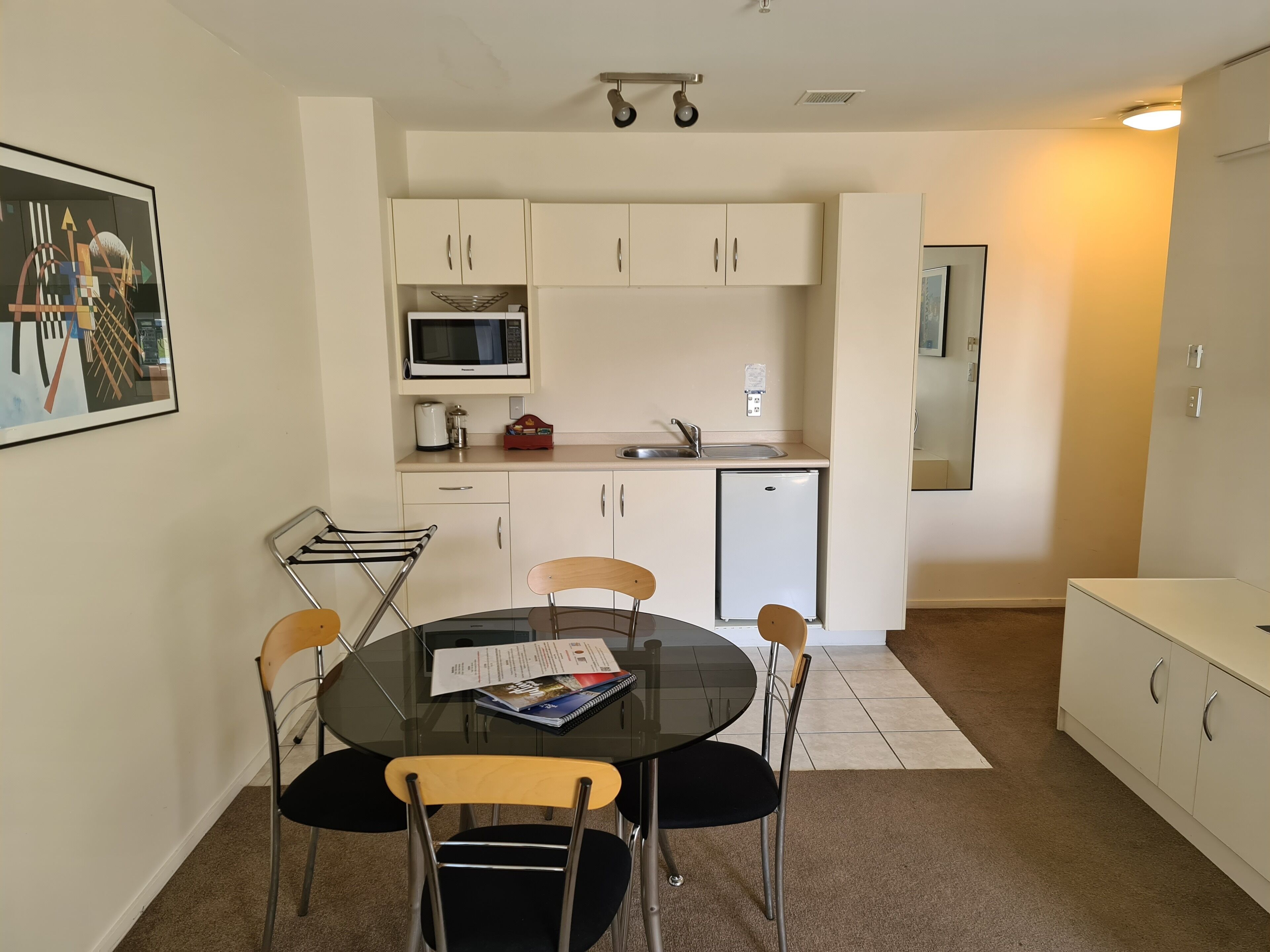 A compact kitchen with white cabinets, a microwave, a sink, and a small refrigerator. There is a dining table with four chairs and a glass top.