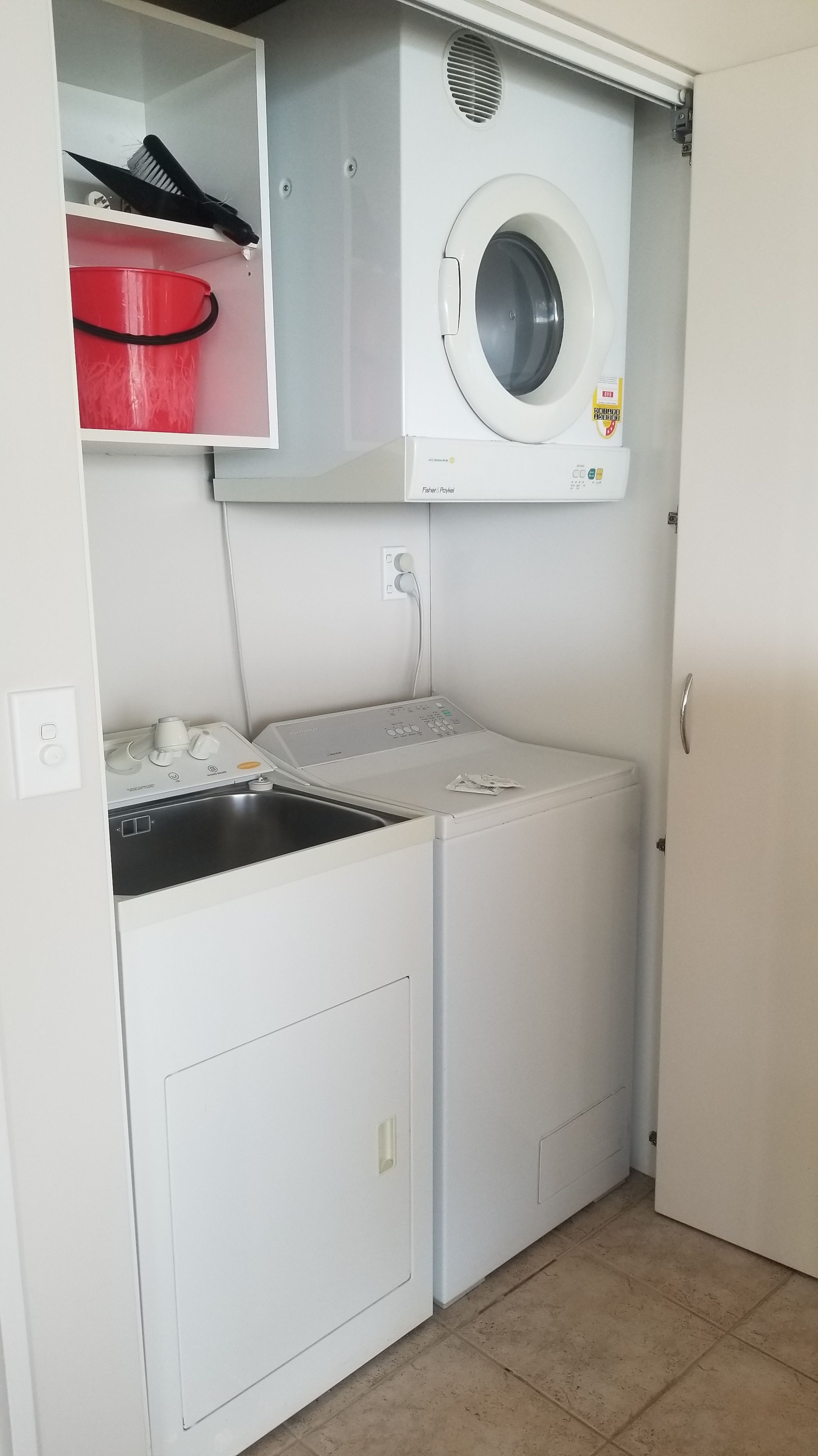 Ett litet och praktiskt tvättrum med en tvättmaskin och en torktumlare som man matar från framsidan, ett handfat och en röd hink.