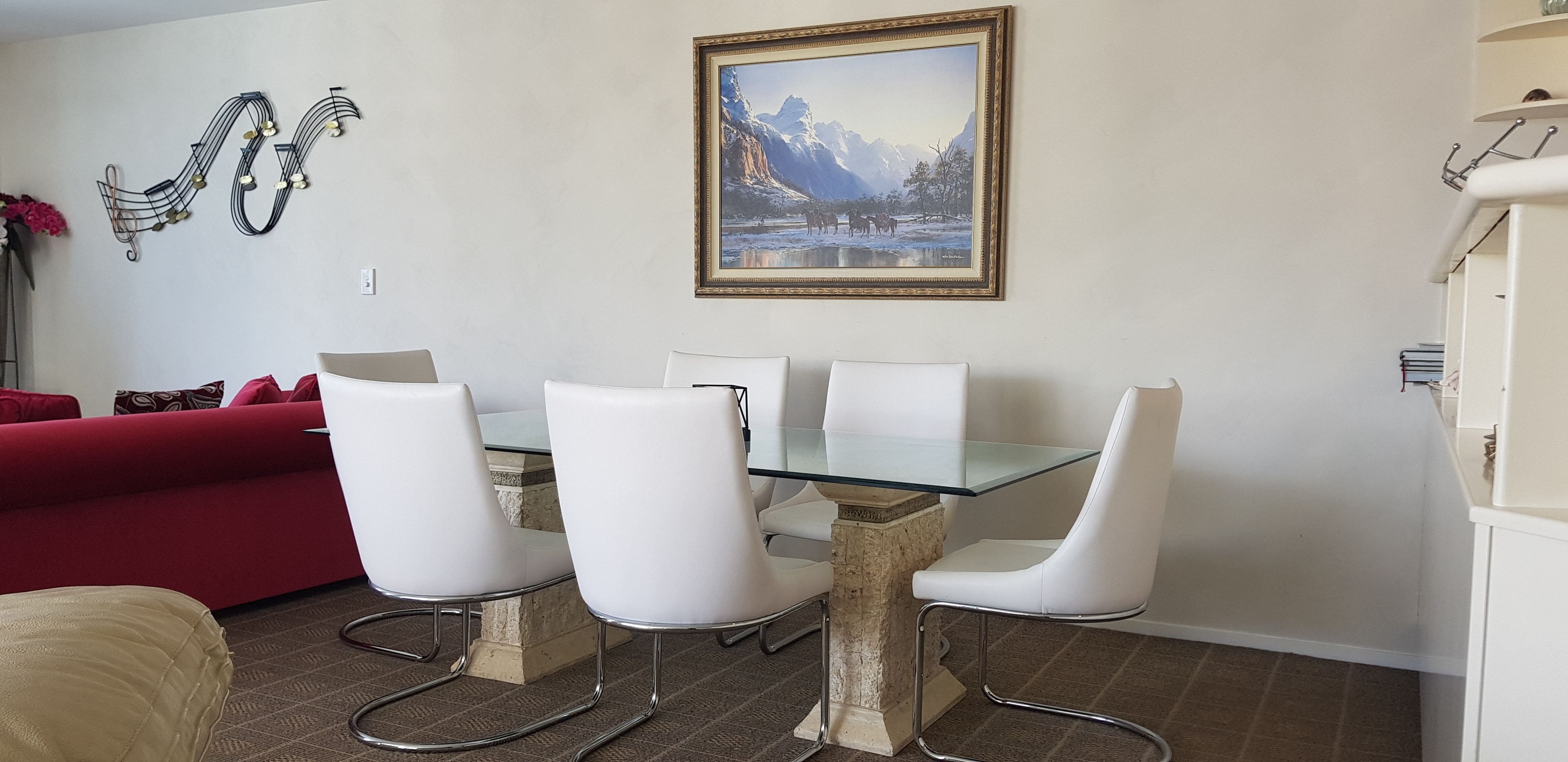 A dining area with a glass-top table and four white chairs. A red couch is visible in the background.