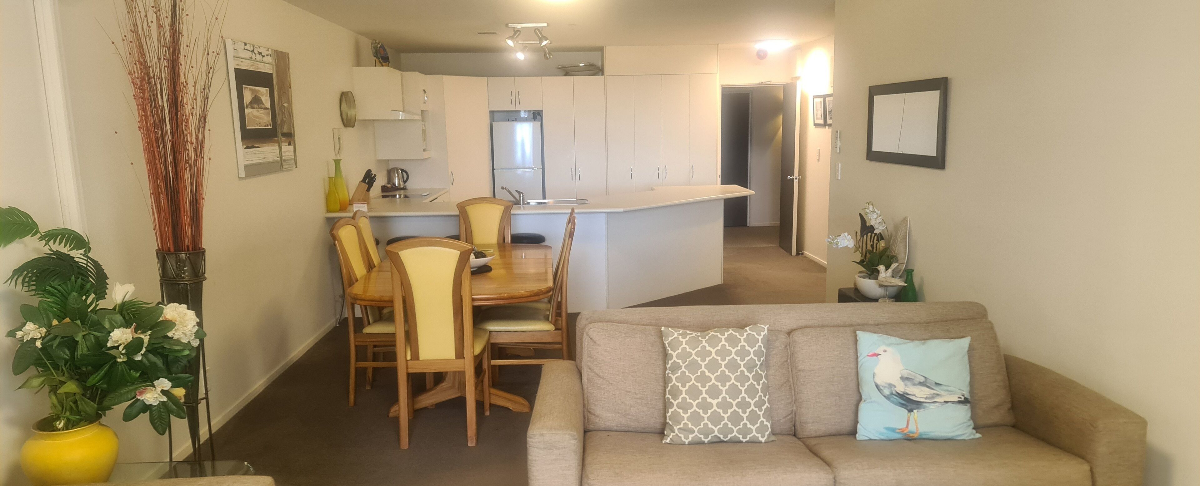 Een moderne keuken met witte kasten, een eethoek met houten stoelen en een beige bank met decoratieve kussens.