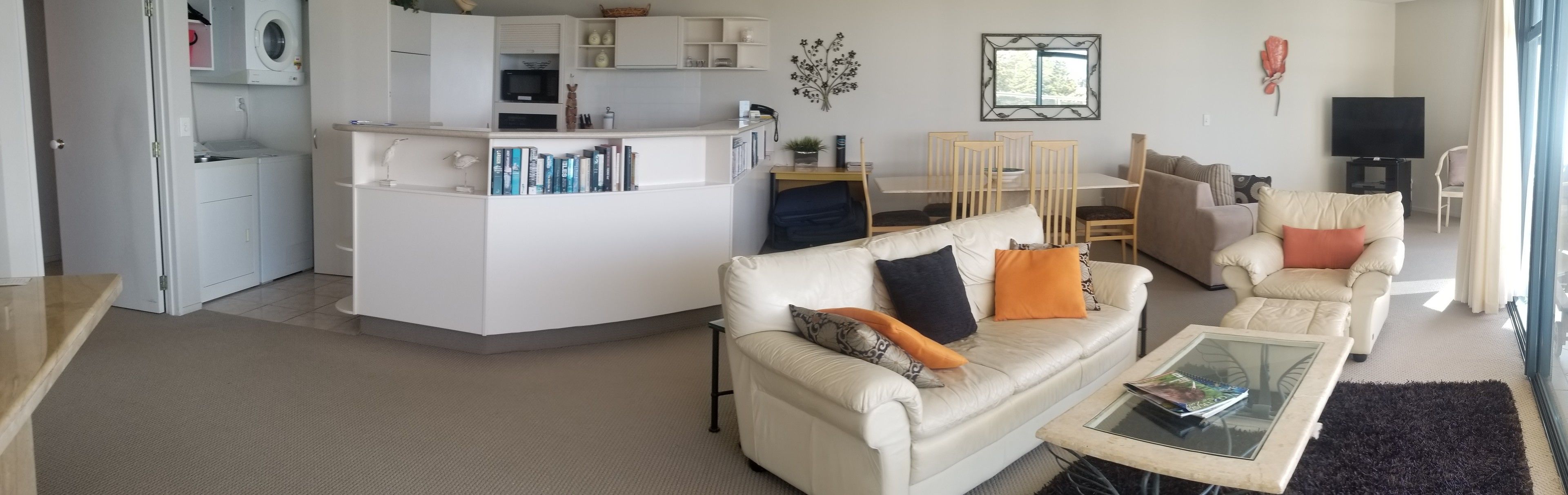 Una sala de estar moderna con un sofá blanco, una mesa de centro de vidrio y una cocina con armarios blancos y un microondas.