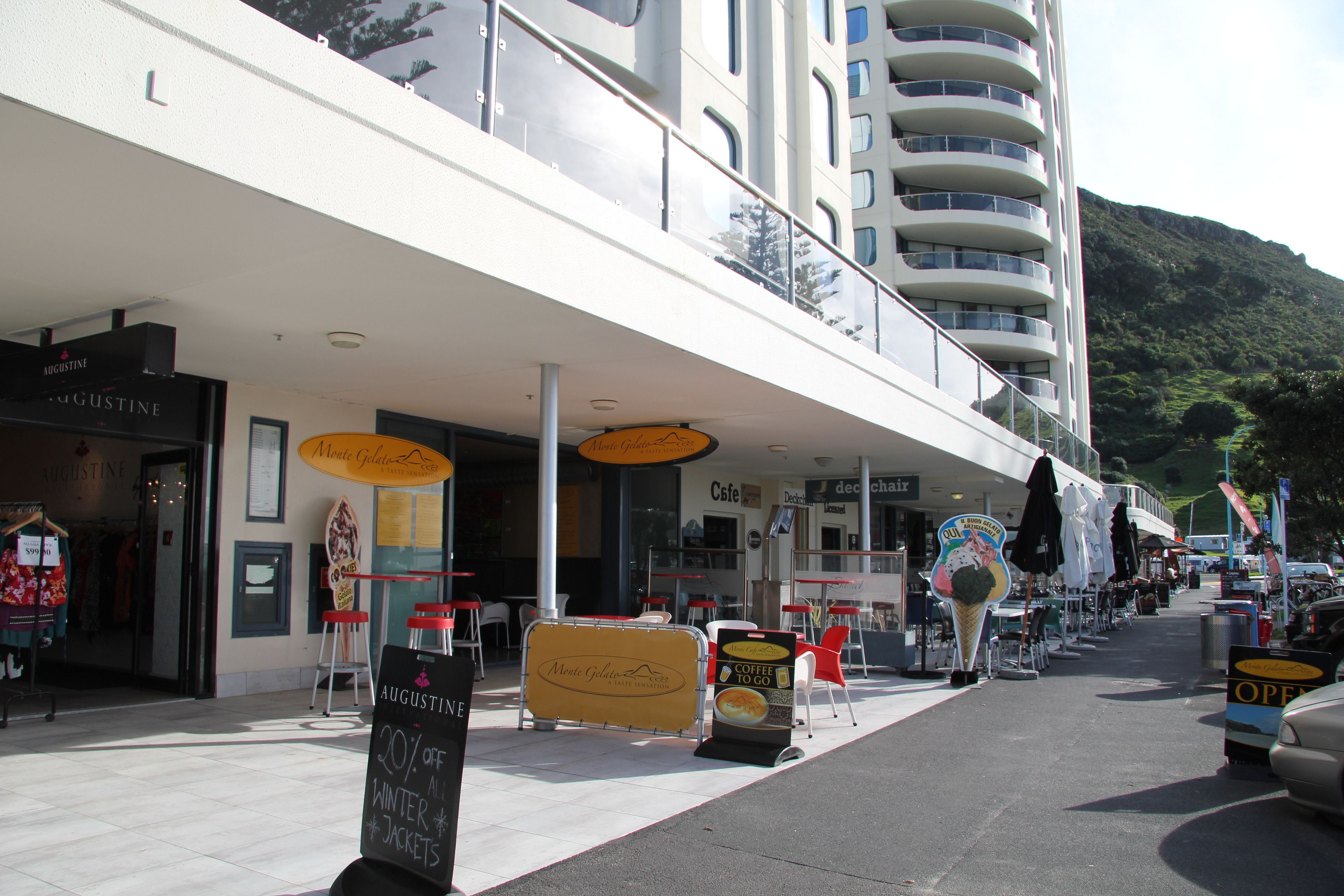 Un edificio moderno con una cafetería y tiendas en la planta baja, mesas al aire libre y vistas a una colina.