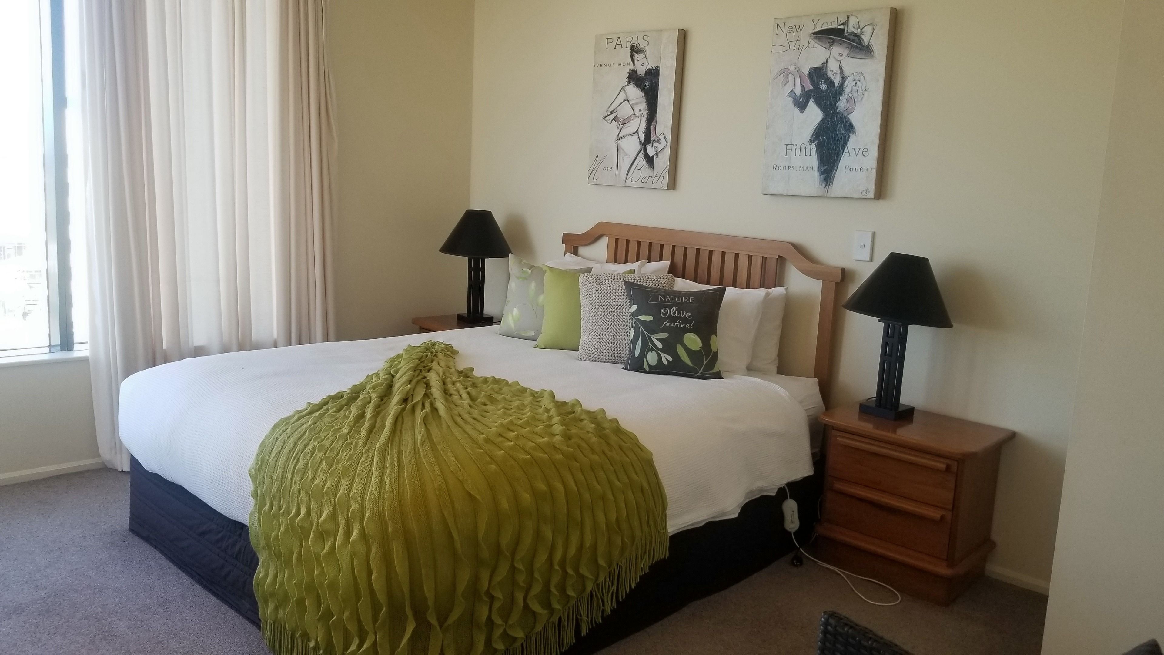 A neatly made bed with a green throw blanket, two bedside lamps, and a wall with two framed artworks.