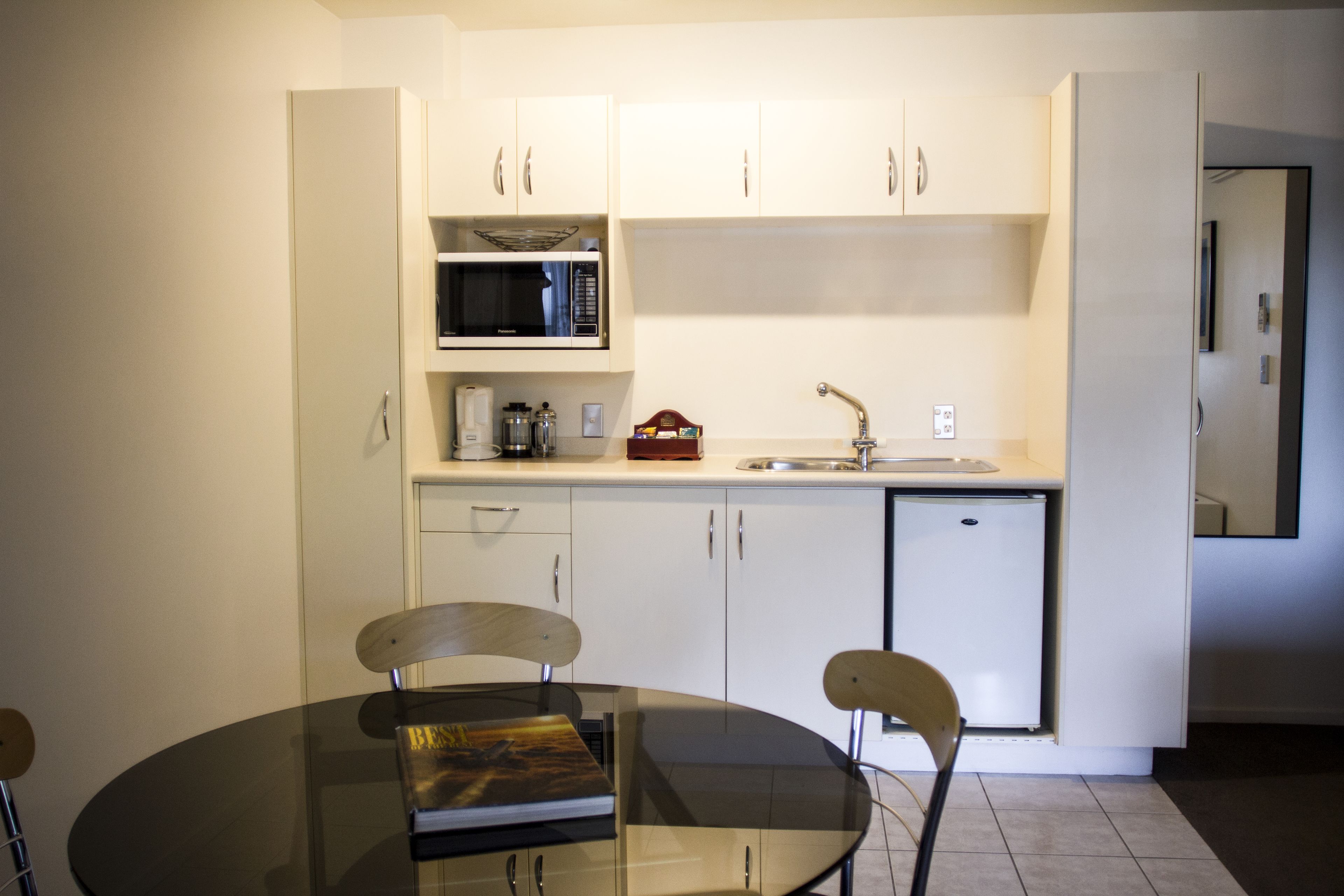 Une cuisine compacte avec des meubles blancs, un four à micro-ondes, un évier et un petit réfrigérateur. On aperçoit une table en verre avec des chaises au premier plan.