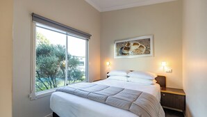 Een slaapkamer met een groot raam, een bed met witte lakens, een nachtkastje en een wandlamp.
