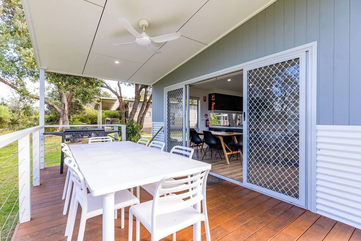 Een terras buiten met een witte tafel en stoelen, een plafondventilator en een schuifdeur naar een keuken.