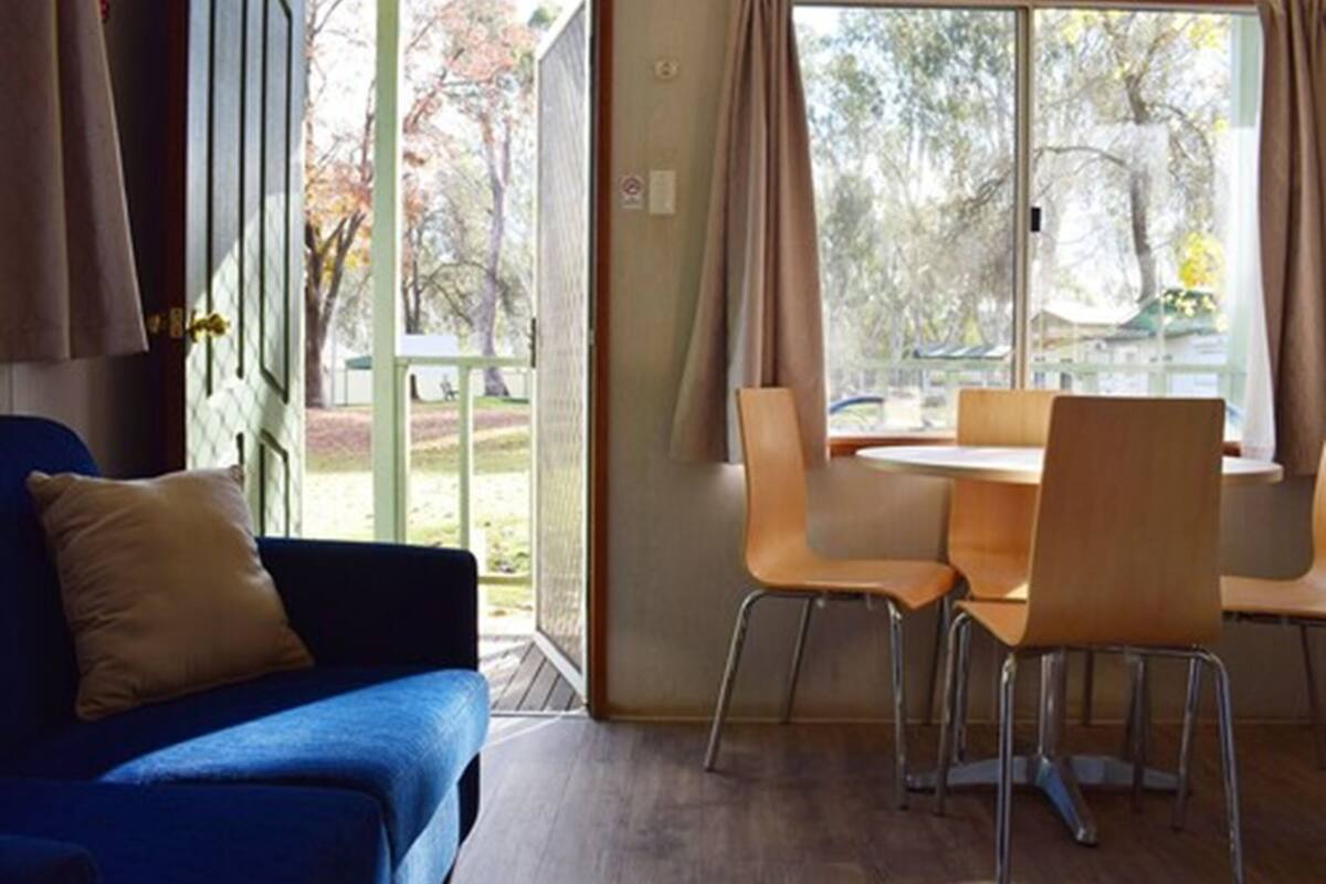 Une pièce avec un canapé bleu, une table à manger et quatre chaises, et une vue sur les arbres par les fenêtres.