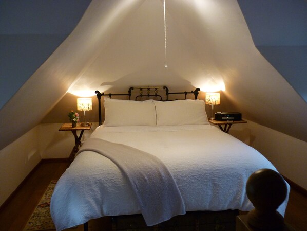 Un dormitorio acogedor con techo triangular, una cama centrada bajo un tejado inclinado, flanqueada por dos mesitas de noche con lámparas.