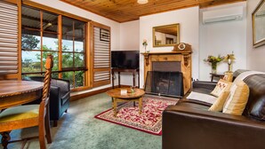 A cozy living room with a fireplace, a television, a dining table with chairs, and a large window offering a view of the outdoors. - Wagners Cottages (Swansea)