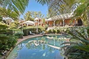A pool surrounded by lush greenery and a large house with a covered patio.