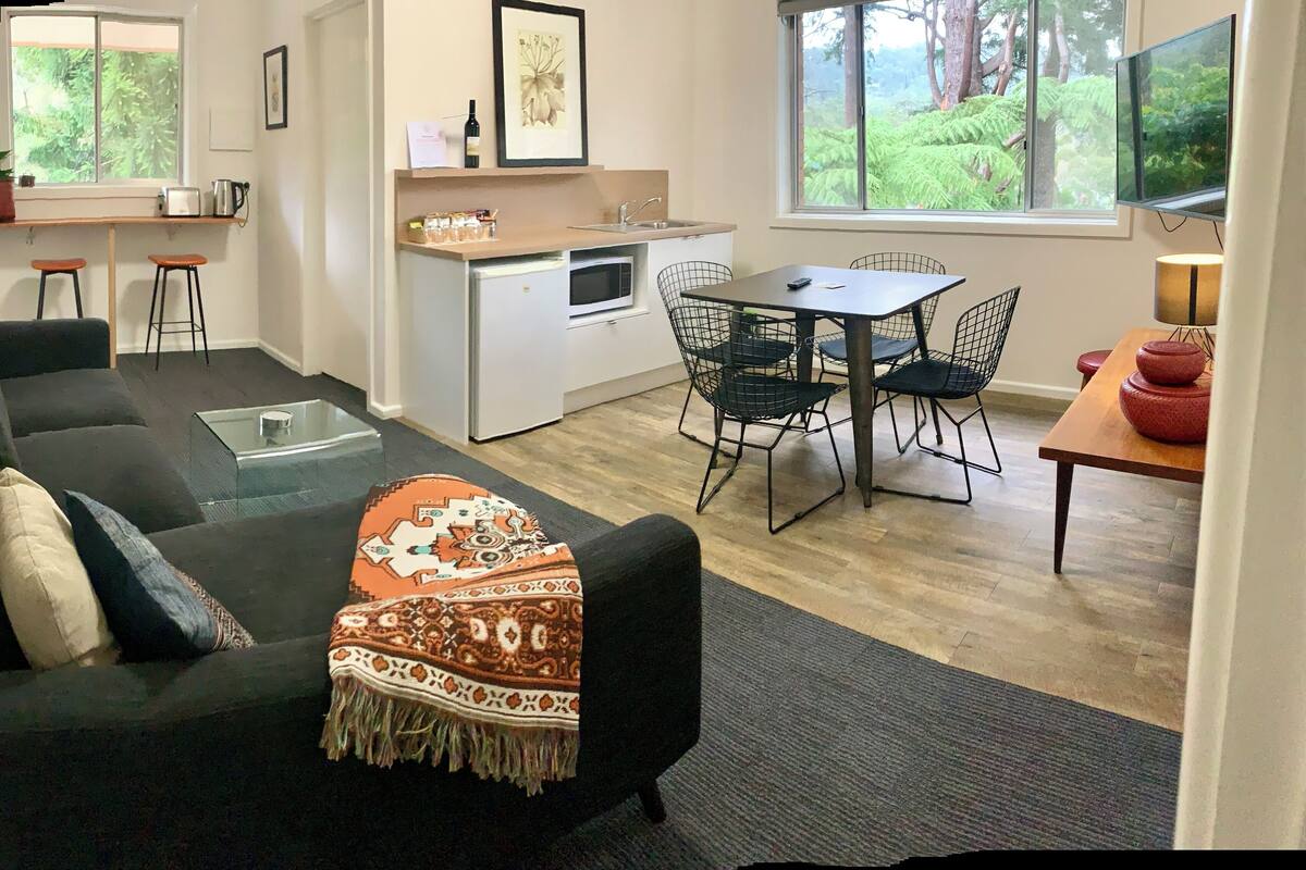 Un salon moderne avec un canapé noir, une table basse en verre et un plaid à motifs. On aperçoit une cuisine équipée d’un four à micro-ondes et un coin repas avec une table et des chaises.