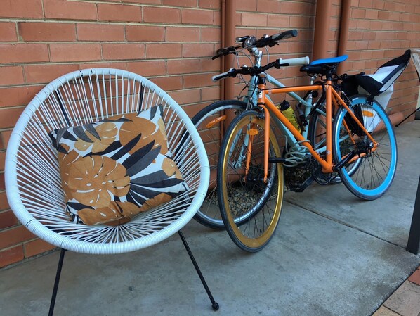 Una silla de mimbre con un cojín estampado, una bicicleta con detalles naranja y azul, y un muro de ladrillo al fondo.