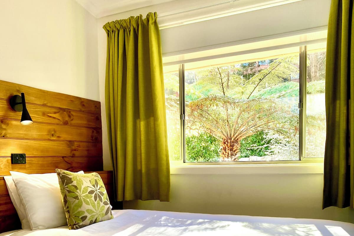 Une chambre à coucher avec une tête de lit en bois, un grand lit recouvert de draps blancs, des rideaux verts et une fenêtre donnant sur un paysage verdoyant.