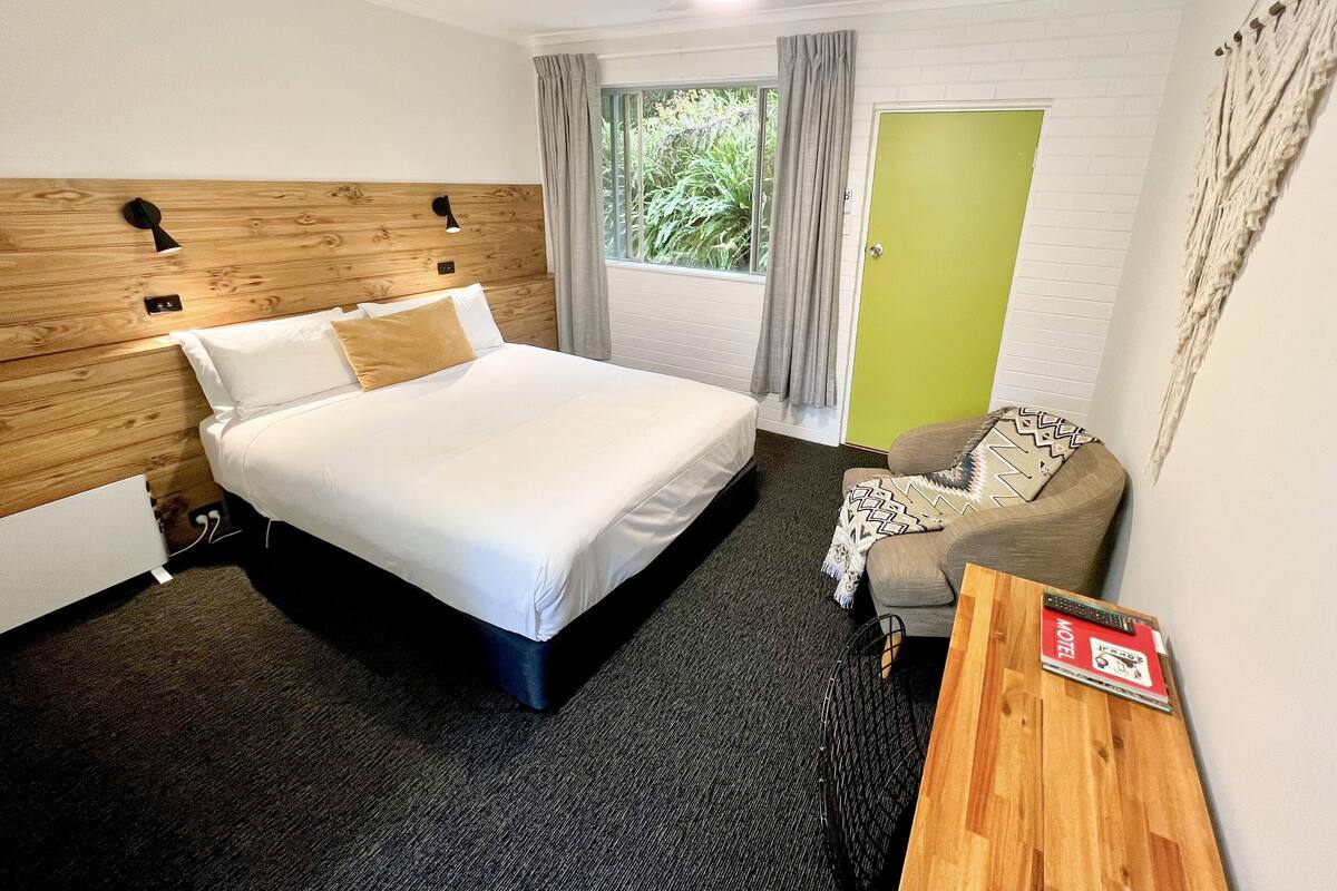 Une chambre d’hôtel avec un lit, une tête de lit en bois, une porte verte, une fenêtre avec des rideaux, un ventilateur de plafond et une petite table sur laquelle est posé un panneau.