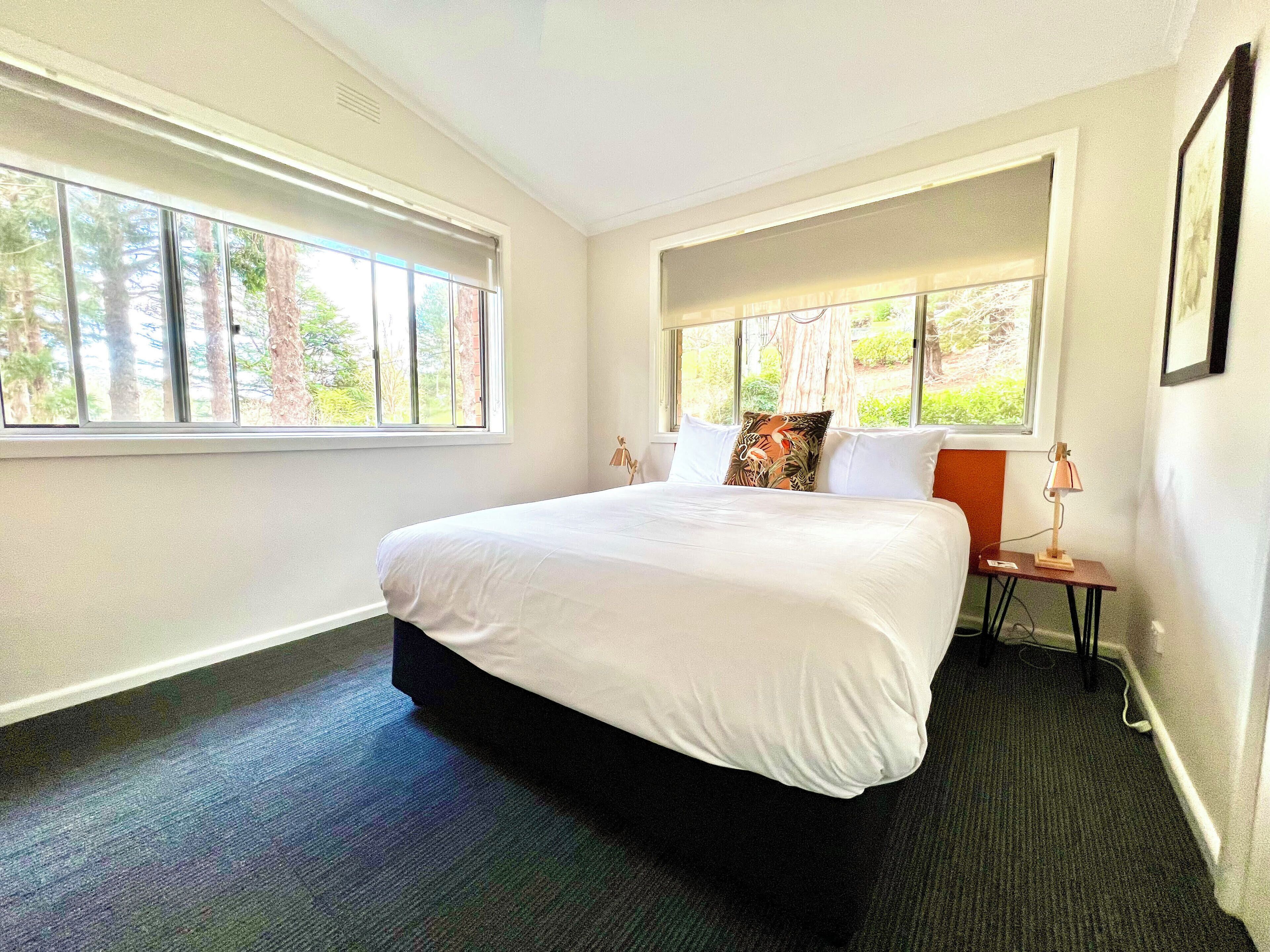A modern hotel room with a large bed, two bedside tables, a nightstand, and a window with a view of trees.