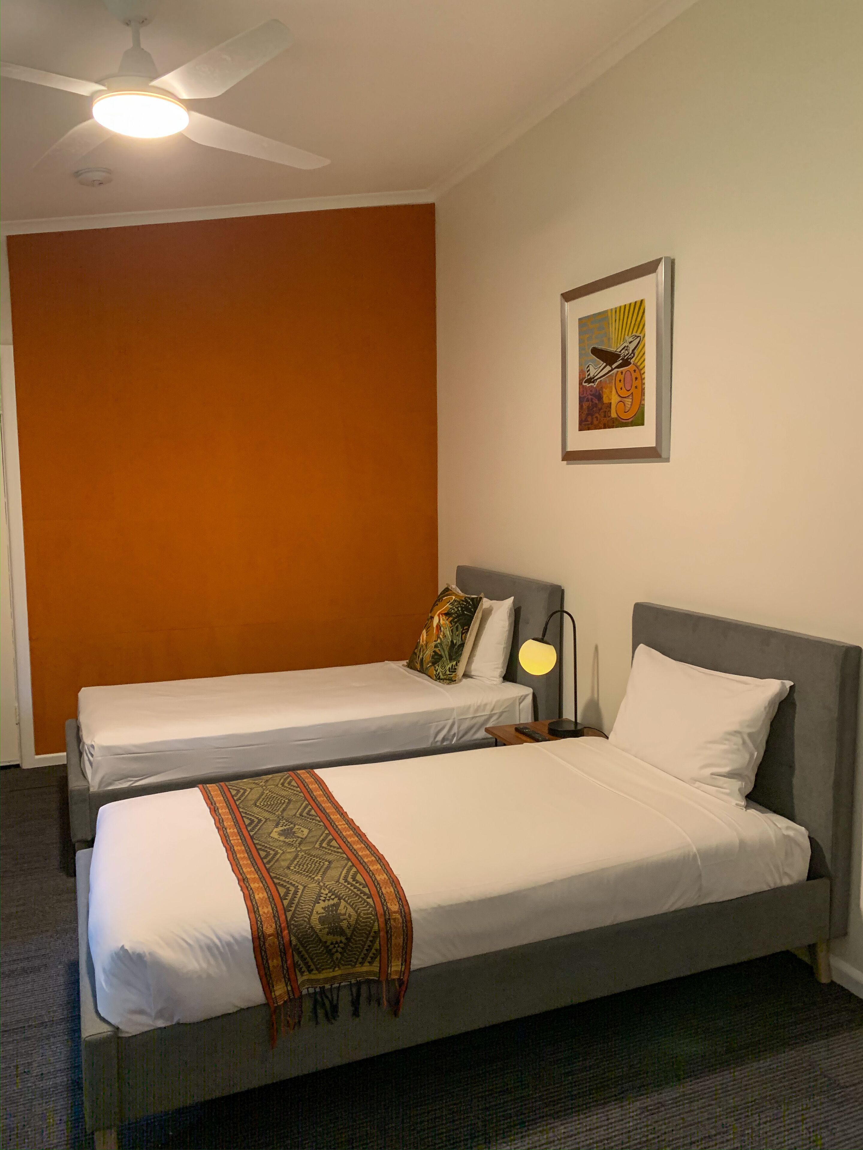 A hotel room with two single beds, a ceiling fan, a framed artwork on the wall, and a carpeted floor.