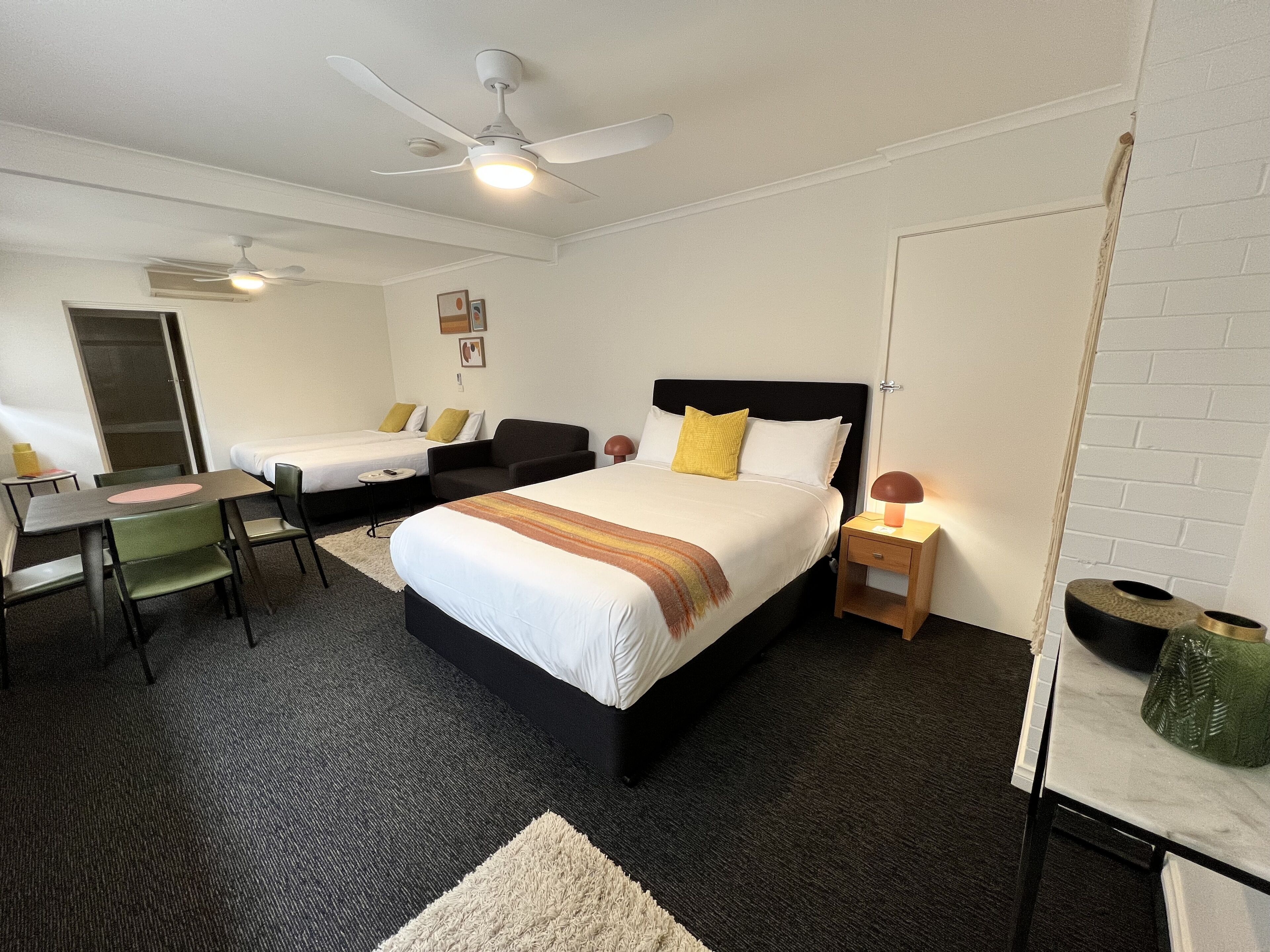 Habitación de hotel con dos camas individuales, un escritorio con sillas, un ventilador de techo y una mesita de noche pequeña con lámpara.