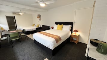 Habitación de hotel con dos camas individuales, un escritorio con sillas, un ventilador de techo y una mesita de noche pequeña con lámpara.