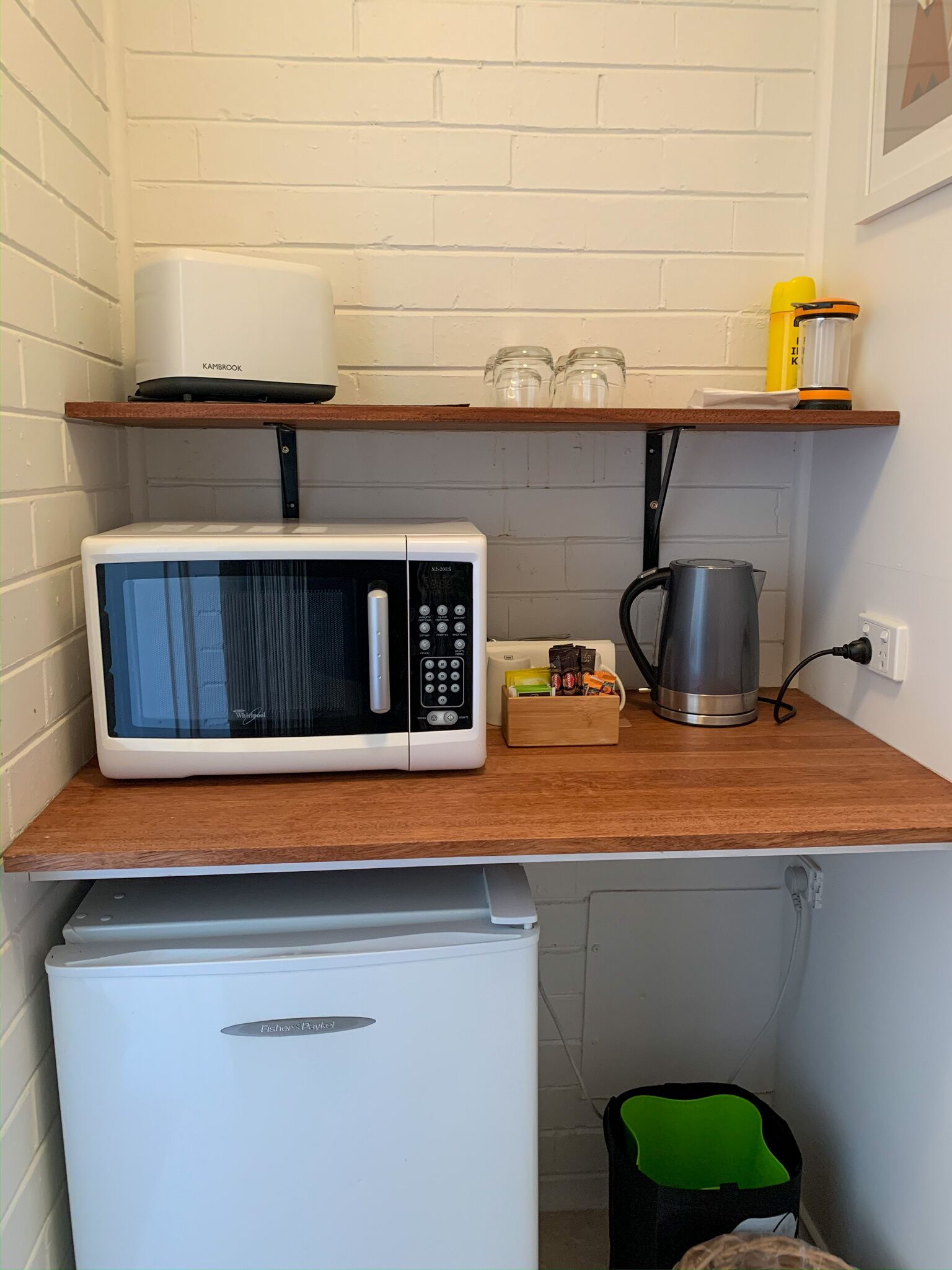 Uma kitchenette compacta com micro-ondas branco, mini geladeira e chaleira sobre bancada de madeira.