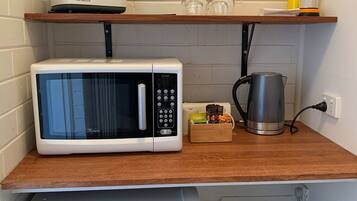 Uma kitchenette compacta com micro-ondas branco, mini geladeira e chaleira sobre bancada de madeira.