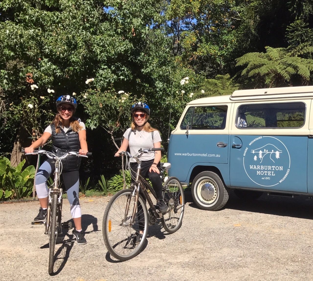 Duas pessoas de bicicleta em frente a uma van com o logotipo do Motel Warburton.