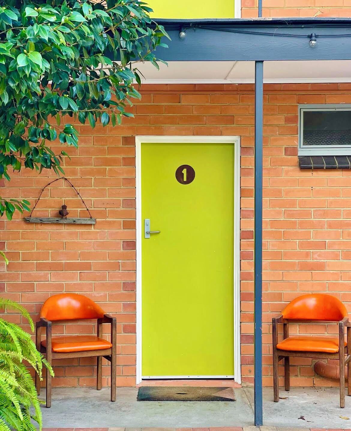 A bright yellow door with a number 1 sign, two orange chairs, and a brick wall.