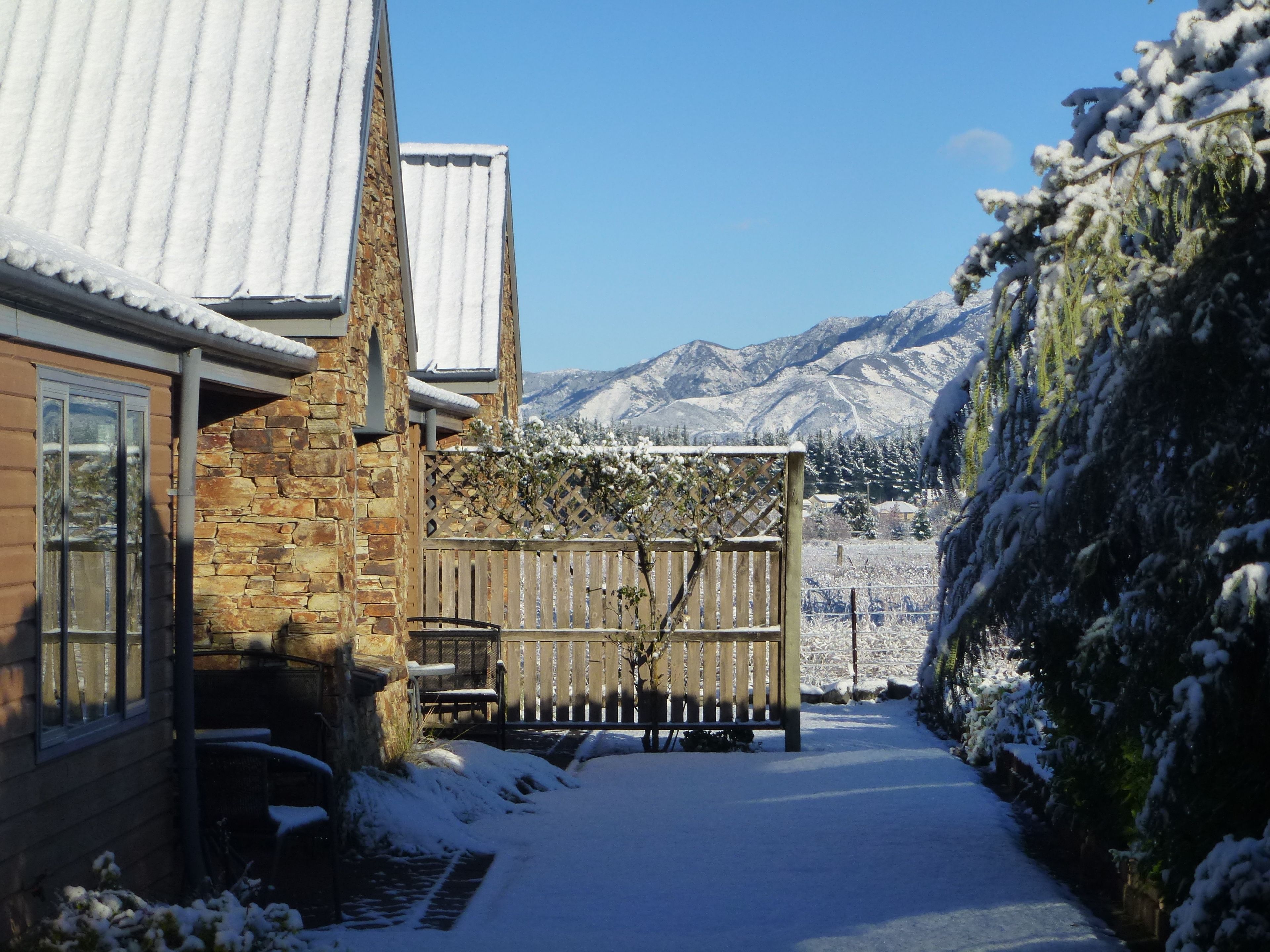 A snow-covered pathway leading to a house with a stone facade and a wooden deck.