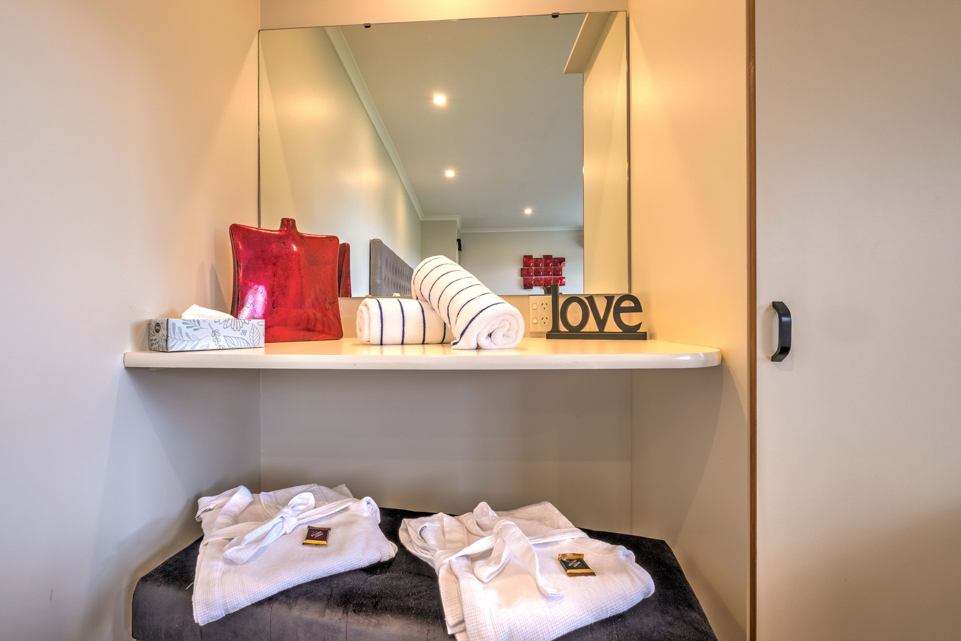 A hotel room with a mirror reflecting a neatly arranged shelf with towels and a decorative sign.