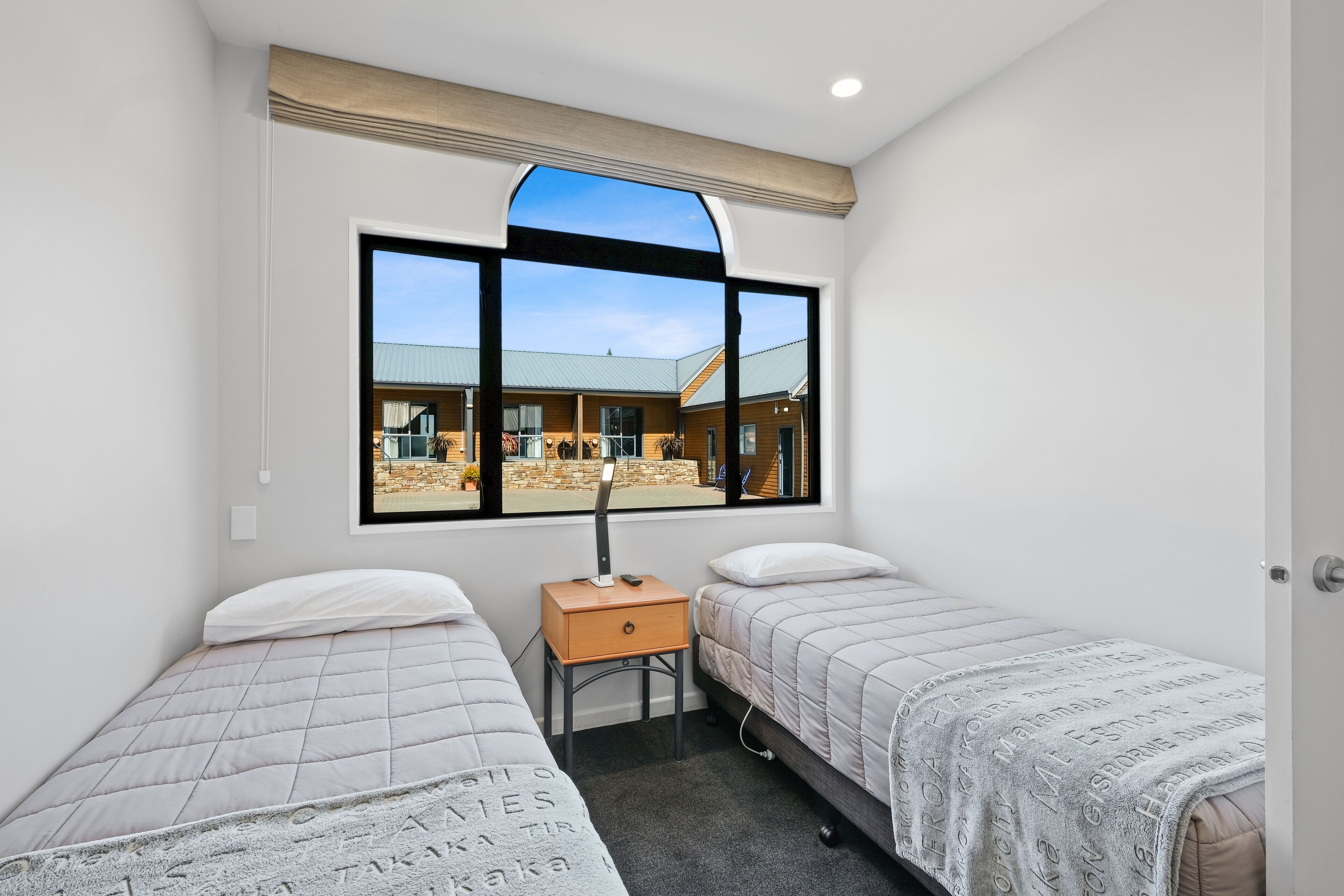 A bedroom with two beds, a window with a view of a building, and a bedside table with a lamp.