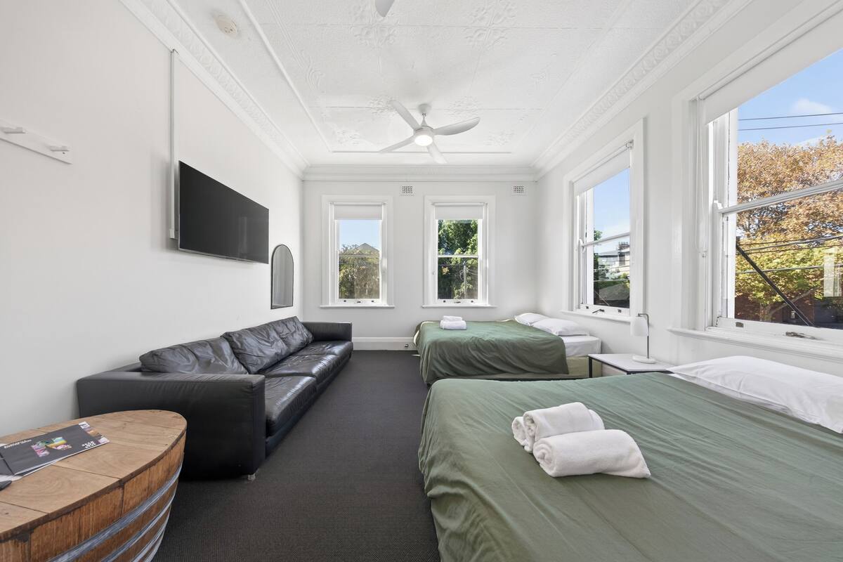 A hotel room with a flat-screen TV, a sofa, two beds, a ceiling fan, and large windows.