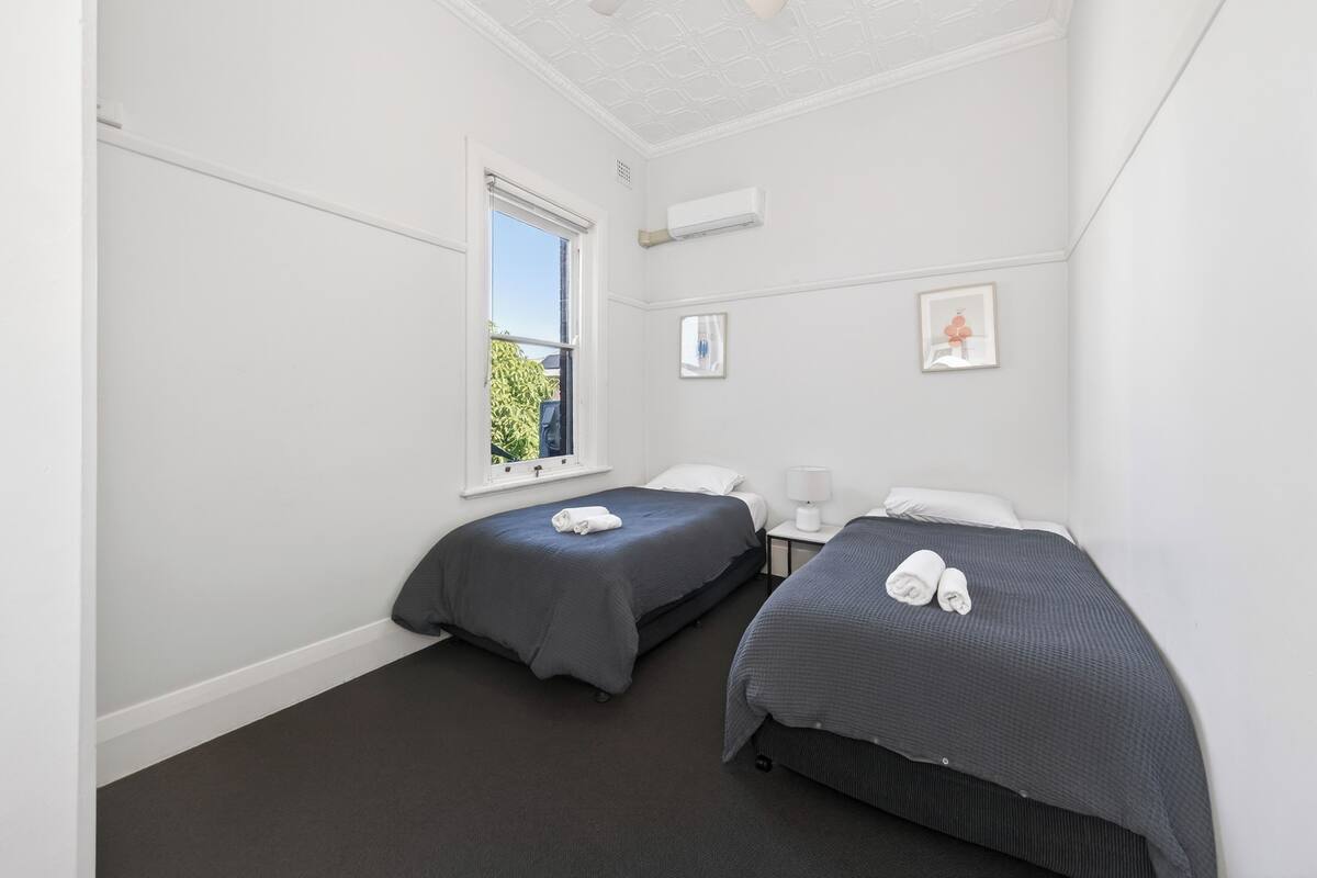 A hotel room with two beds, a window, a nightstand, and a wall-mounted air conditioner.