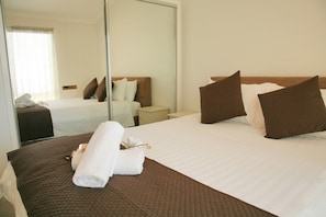 A hotel room with a large bed, a bedside table, a mirror, and a window with blinds. - Surf Beach Holiday Park (Kiama)