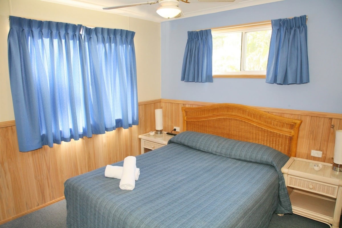 Une chambre d’hôtel comprenant un lit, deux tables de chevet, une commode, une fenêtre avec des rideaux et un ventilateur de plafond.