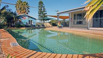 A swimming pool with a brick border, surrounded by palm trees and a building with a large window.