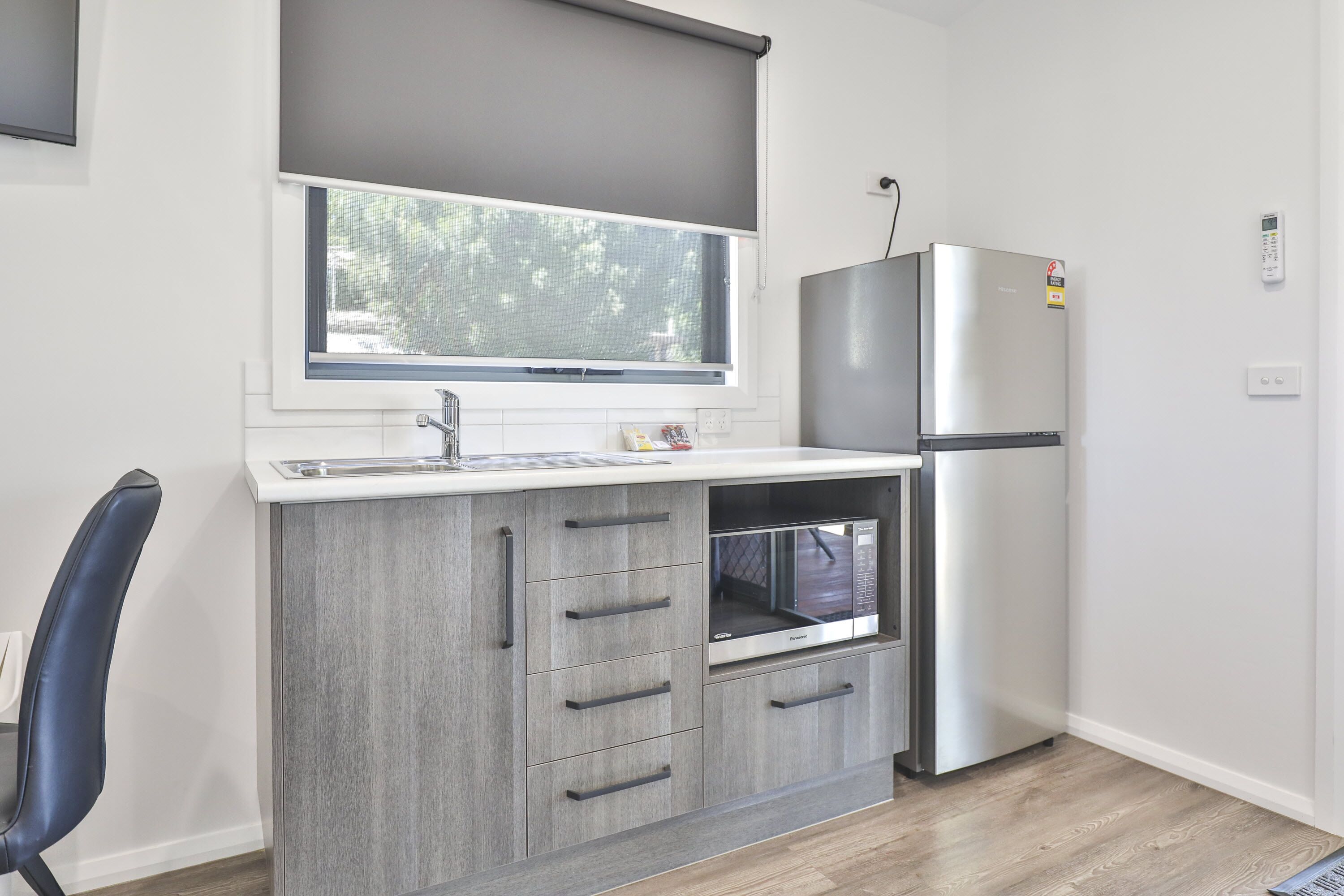 A compact kitchen with a built-in microwave, a stainless steel refrigerator, and a window with a roller blind.