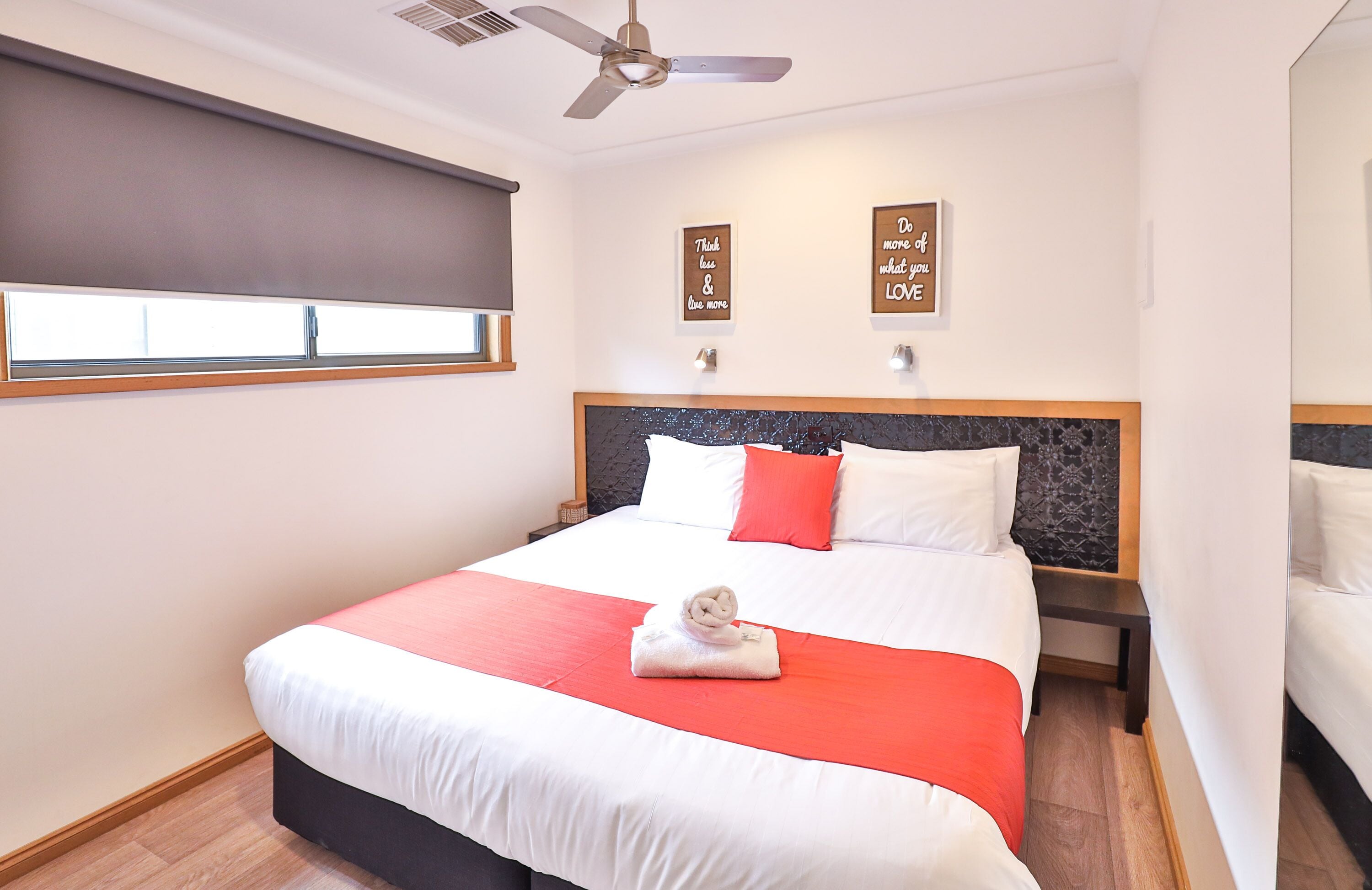 A neatly made bed with a red and white bedspread, a folded towel, and two wall-mounted light fixtures.