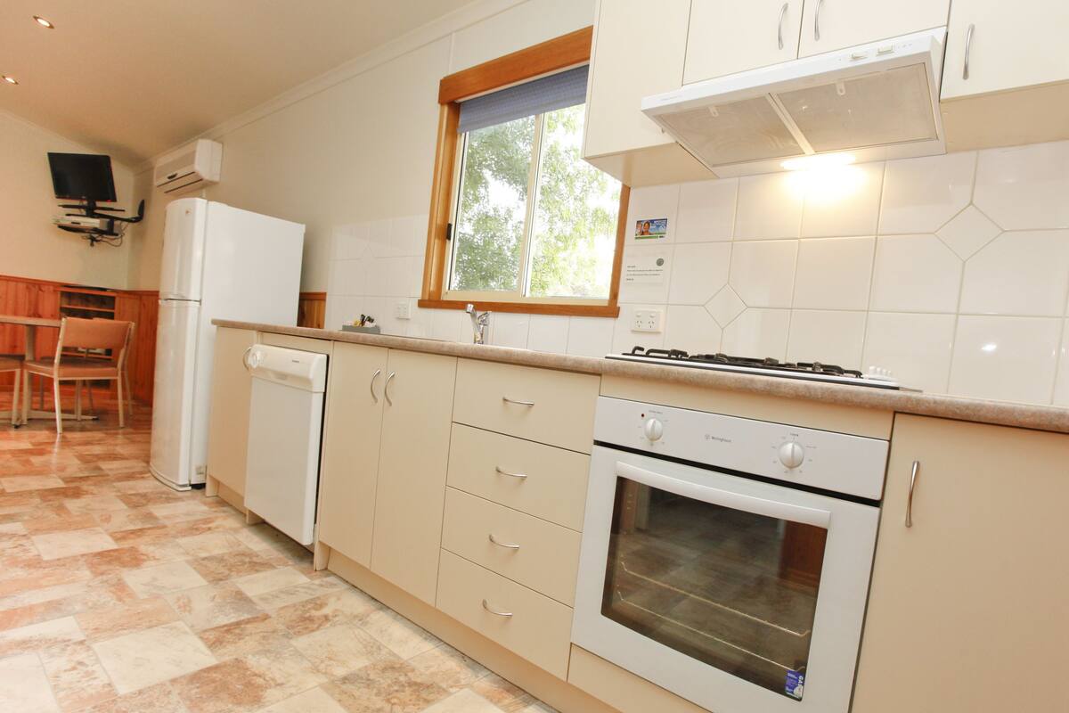 A kitchen with a dishwasher, oven, and stove. There is a window above the sink and a television mounted on the wall.