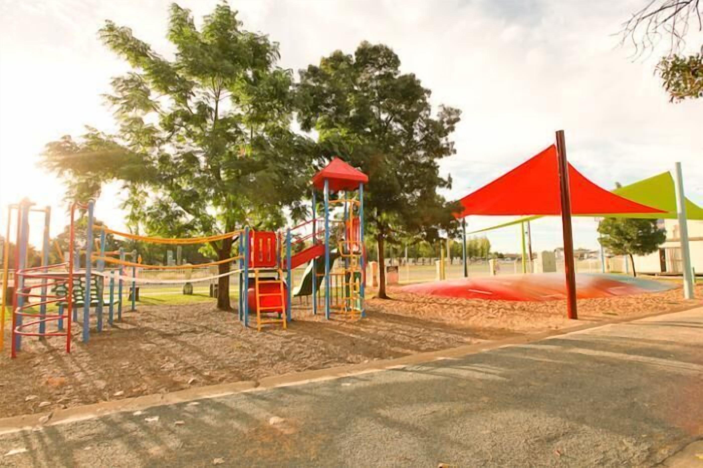 Un parque infantil con diversas estructuras de juego, incluyendo toboganes y juegos de escalar, rodeado de árboles y cubierto por un toldo rojo.